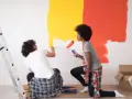 Un couple peint un mur en jaune et orange. Ils utilisent des rouleaux pour nettoyer peinture glycero. Une échelle est à gauche.