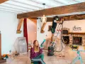 Un homme ponce une poutre en bois avec une ponceuse, tandis qu'une femme tient des outils.