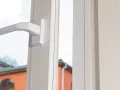 Poignée blanche d'une fenêtre, révélant le mécanisme cremone porte fenetre. Dehors, des maisons et des arbres verts.