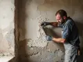 Un homme enlève un crépi intérieur abîmé avec une spatule. Il porte des gants et une salopette.