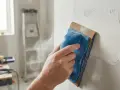 Main d'un artisan ponçant un mur en placo avec un grain adapté pour un fini lisse. La poussière s'envole.