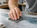 Main d'artisan ponçant une surface pour peindre sur du vernis. La poussière fine indique le travail de préparation.