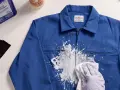 Une main gantée essaie de nettoyer peinture acrylique sèche sur une veste bleue avec un chiffon. Un grattoir et un produit sont à proximité.