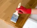 Application d'huile de lin sur un plateau en bois de douglas. La main gantée de rouge applique le produit avec un pinceau.