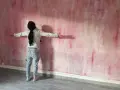 Une personne admire une peinture à la spatule sur mur aux tons roses et blancs, les bras écartés.