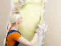Une femme en salopette bleue et t-shirt orange mesure un mur pour le détapisser puis peindre.