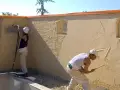 Deux ouvriers appliquent du crépi extérieur. L'un utilise un pistolet pour projeter le matériau, l'autre lisse le crépi à la truelle.