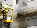 Un ouvrier en nacelle jaune utilise un nettoyeur haute pression pour décaper la peinture d'une façade en pierre, créant un nuage de vapeur.