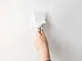 Une main utilise un couteau à mastic pour enlever de la vieille peinture écaillée d'un mur blanc, une étape essentielle pour savoir comment enlever la colle sur un mur avant de peindre.