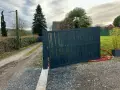 Portail en pente avec gonds sur mesure. Le portail gris foncé est ouvert, révélant un chemin de gravier et une allée bordée de haies.
