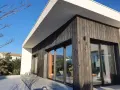 Façade moderne avec bardage en **bois brulé**, grandes baies vitrées reflétant le ciel bleu et la végétation.