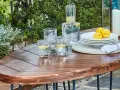 Table en bois couleur acacia, prête pour un apéritif en plein air avec des verres d'eau citronnée, des citrons et une bougie.