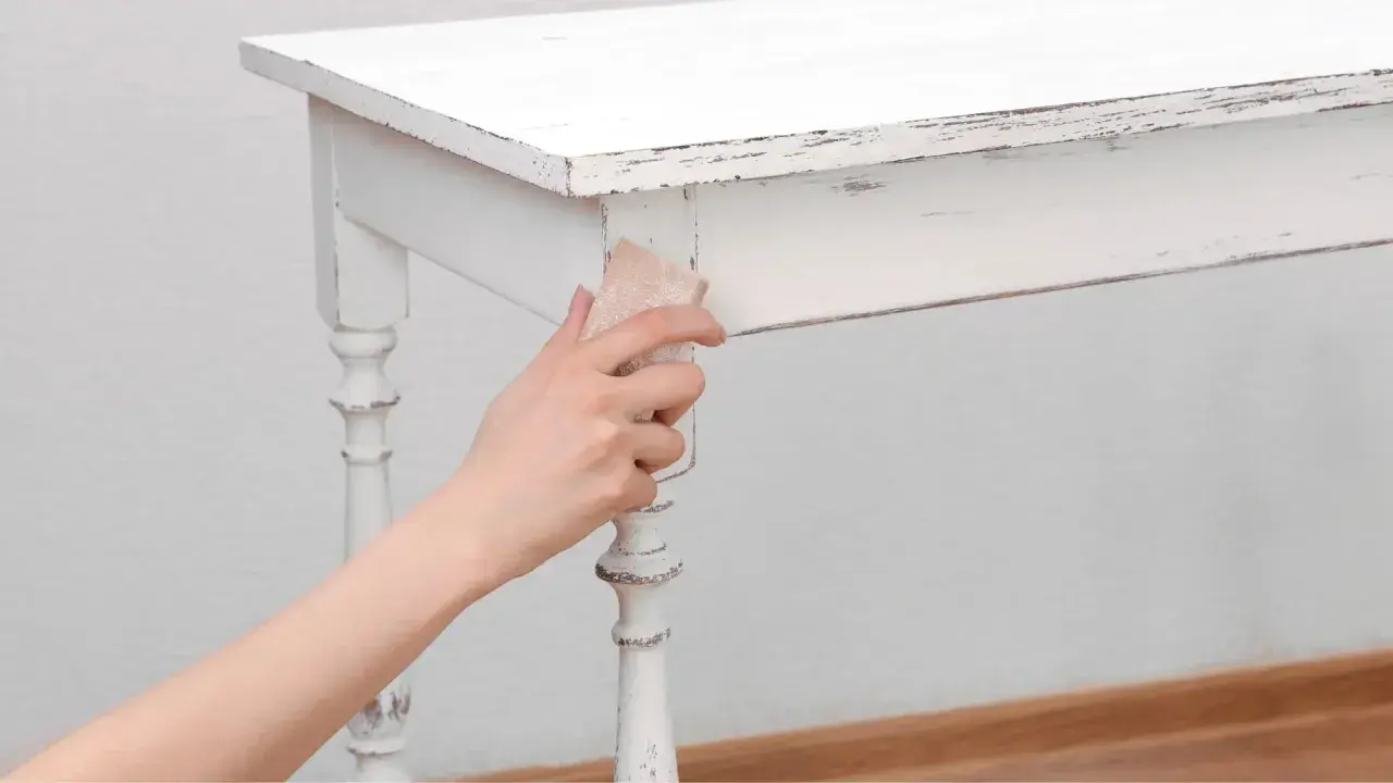 Main tenant enlever peinture sur bois d'une table blanche vieillie, révélant le grain du bois.