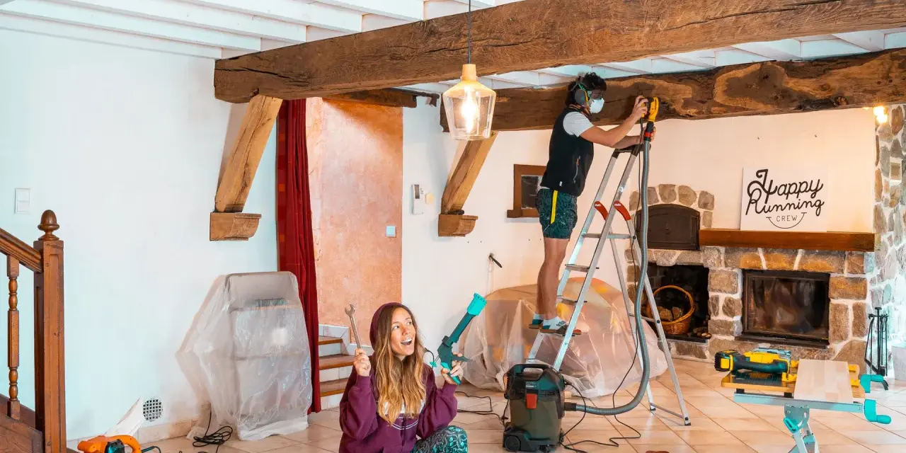 Un homme ponce une poutre en bois avec une ponceuse, tandis qu'une femme tient des outils.