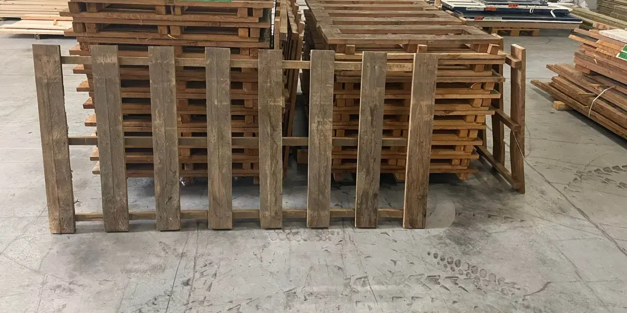 Des planches de bois recycle, issues de palettes, forment une barrière devant d'autres palettes empilées.