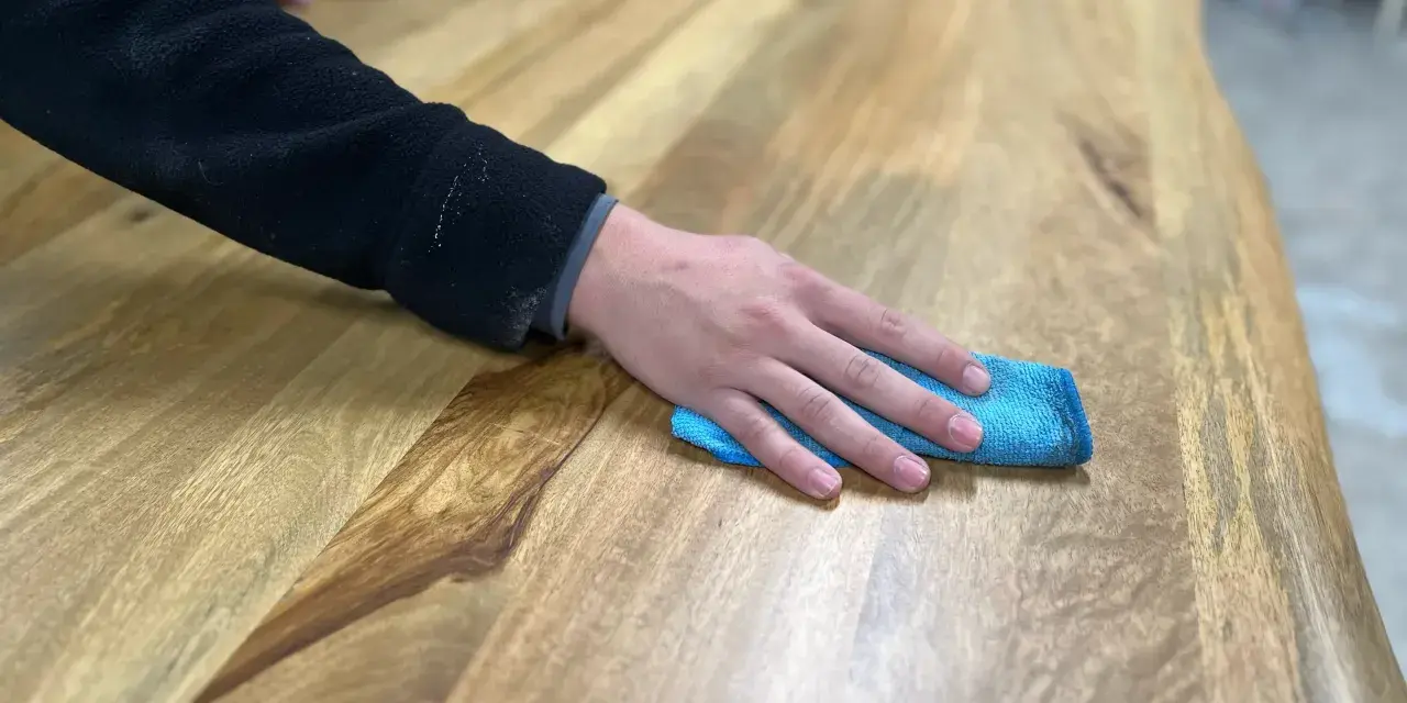 Main d'une personne passant un chiffon bleu sur une table en bois, montrant comment nettoyer une table en bois.