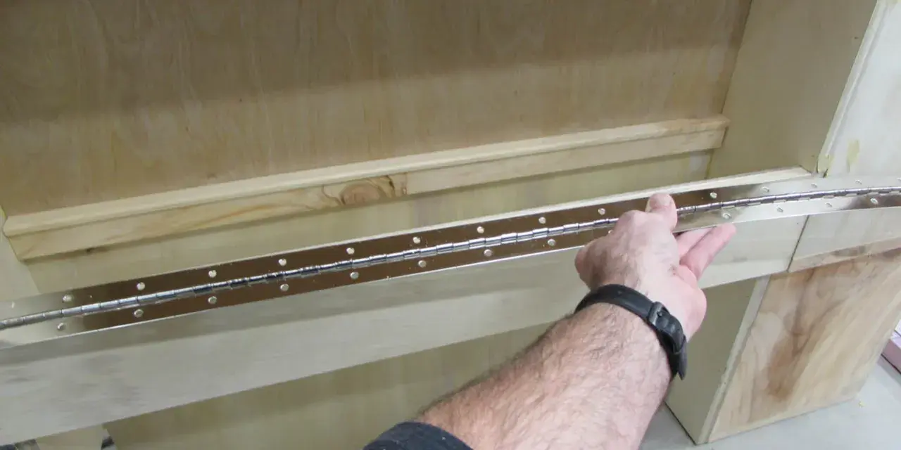 Main tenant une longue charnière piano pour la fixer sur un meuble en bois.