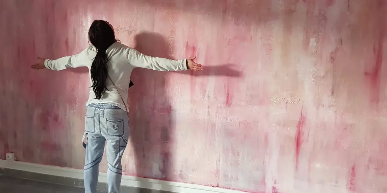 Une personne admire une peinture à la spatule sur mur aux tons roses et blancs, les bras écartés.