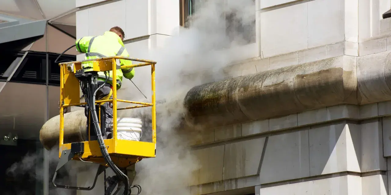 Un ouvrier en nacelle jaune utilise un nettoyeur haute pression pour décaper la peinture d'une façade en pierre, créant un nuage de vapeur.