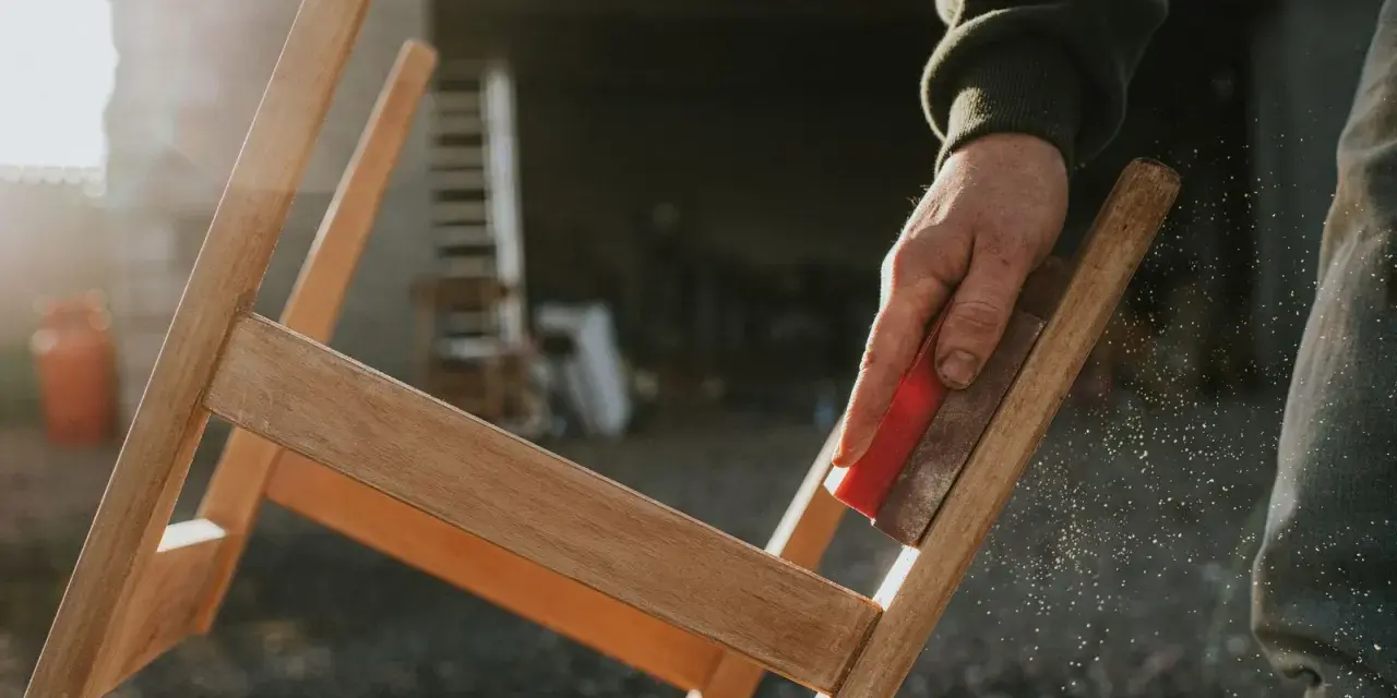 Main d'artisan ponçant un meuble en bois, préparant la surface pour enlever peinture sur bois acétone. Poussière de bois en suspension.