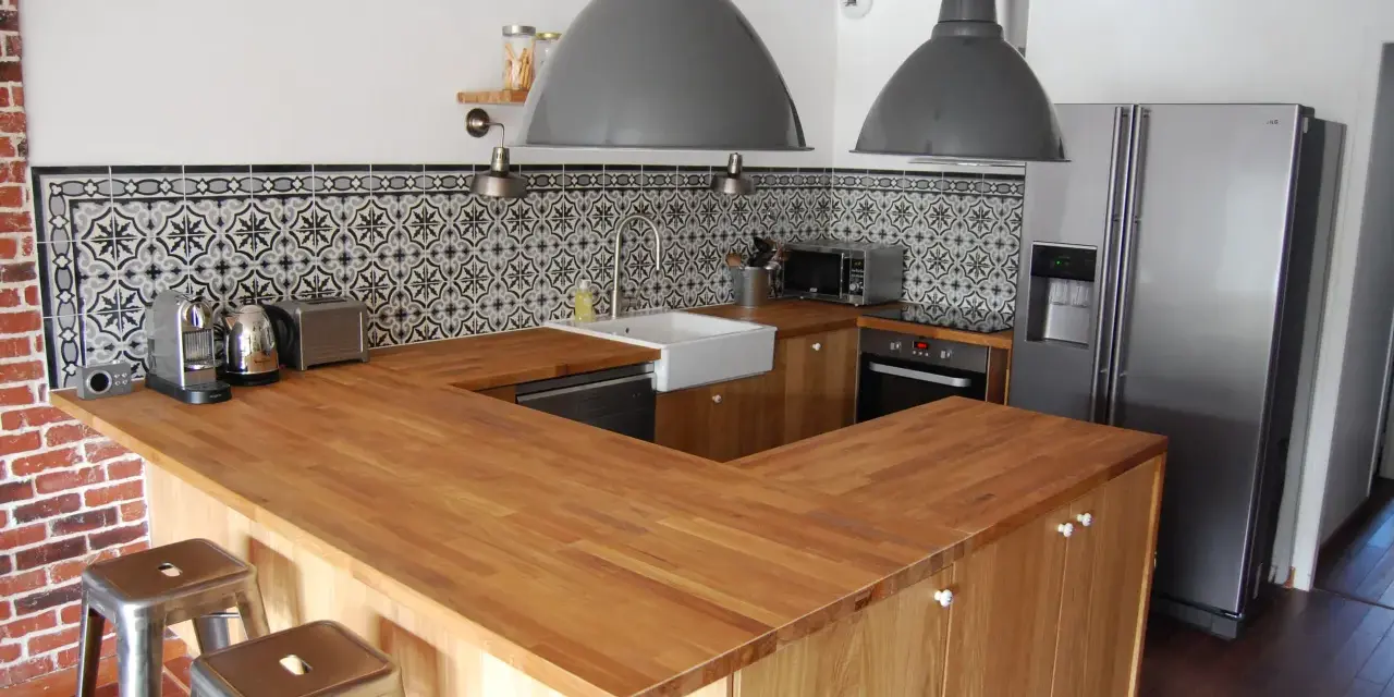 Cuisine moderne avec îlot central en bois, carrelage mural graphique et tabourets industriels. On dirait qu'on a réussi à faire une cuisine avec des vieux meubles revisités.