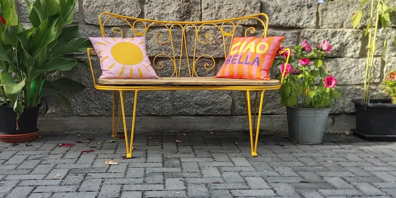 Banc en fer forgé jaune vif, repeint, avec coussins décoratifs. Un soleil rose et "Ciao Bella" sur fond rayé.