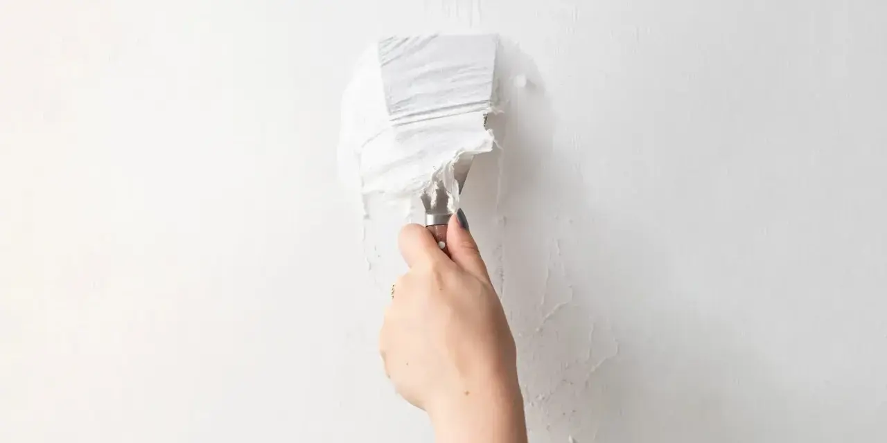 Une main utilise un couteau à mastic pour enlever de la vieille peinture écaillée d'un mur blanc, une étape essentielle pour savoir comment enlever la colle sur un mur avant de peindre.