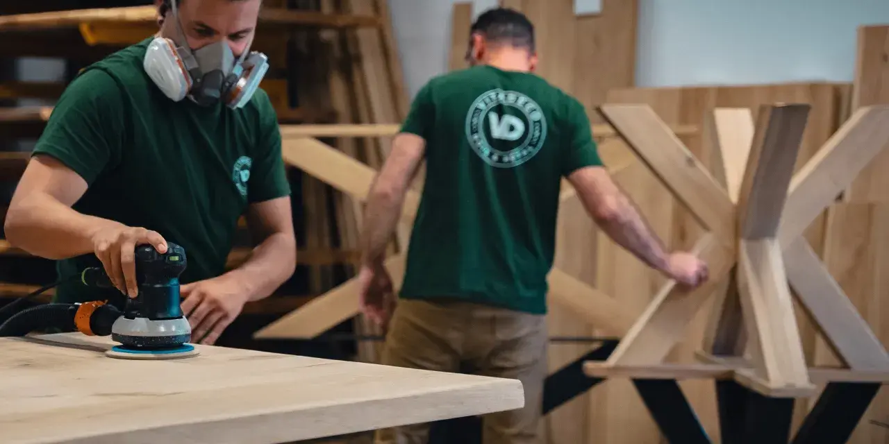 Un artisan, masqué, utilise une ponceuse pour poncer une table en bois. Un autre artisan travaille sur un pied de table en arrière-plan.