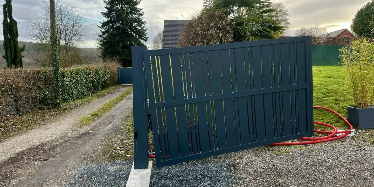 Portail en pente avec gonds sur mesure. Le portail gris foncé est ouvert, révélant un chemin de gravier et une allée bordée de haies.