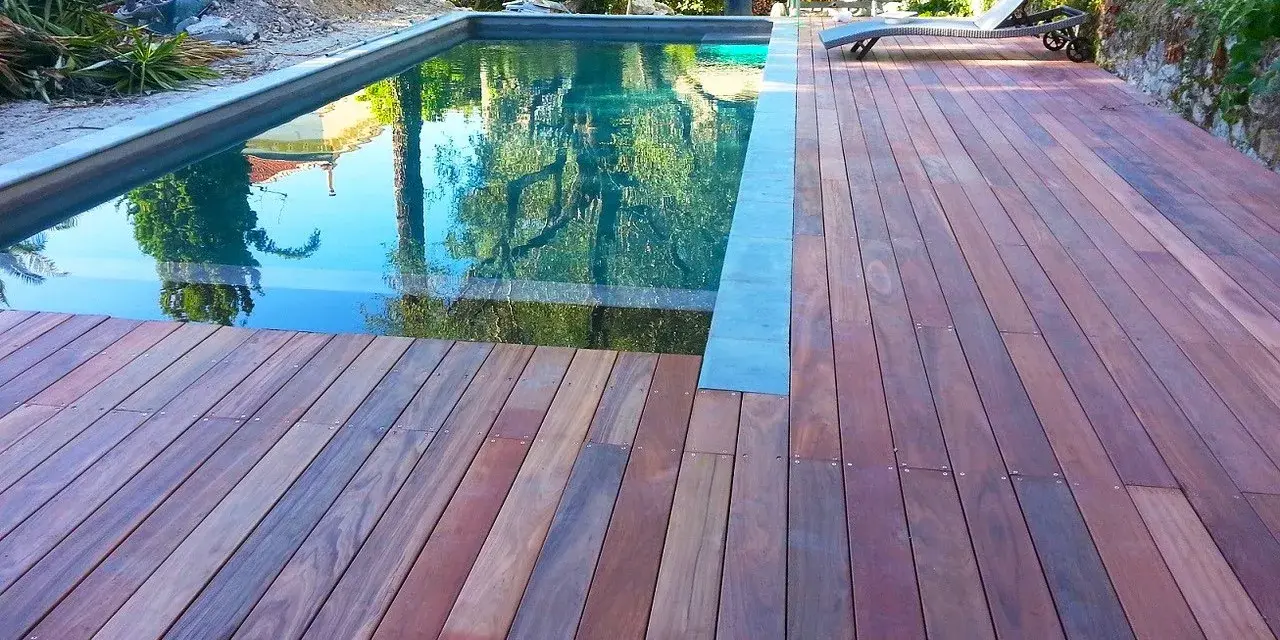 Terrasse en padouk vieilissant, reflétant les arbres dans la piscine. Un transat attend au soleil.