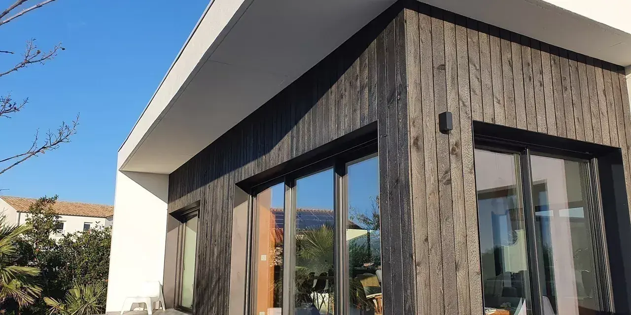 Façade moderne avec bardage en **bois brulé**, grandes baies vitrées reflétant le ciel bleu et la végétation.
