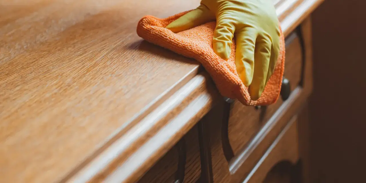 Main d'une personne portant un gant jaune, essuyant un meuble en bois avec un chiffon orange. La qualité du bois est mise en valeur par la lumière.