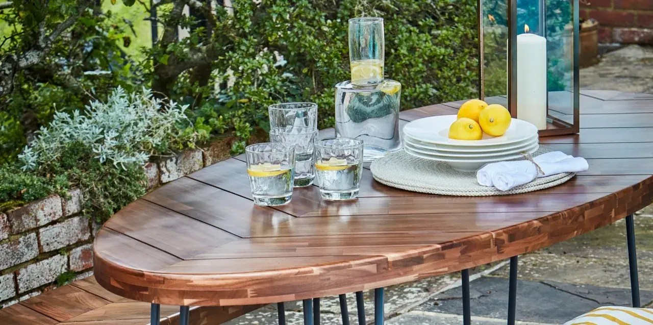 Table en bois couleur acacia, prête pour un apéritif en plein air avec des verres d'eau citronnée, des citrons et une bougie.