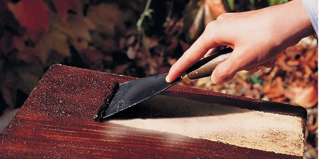 Main d'une personne utilisant un grattoir pour enlever de la peinture d'un meuble en bois, comme avec un décapant peinture bois fait maison.