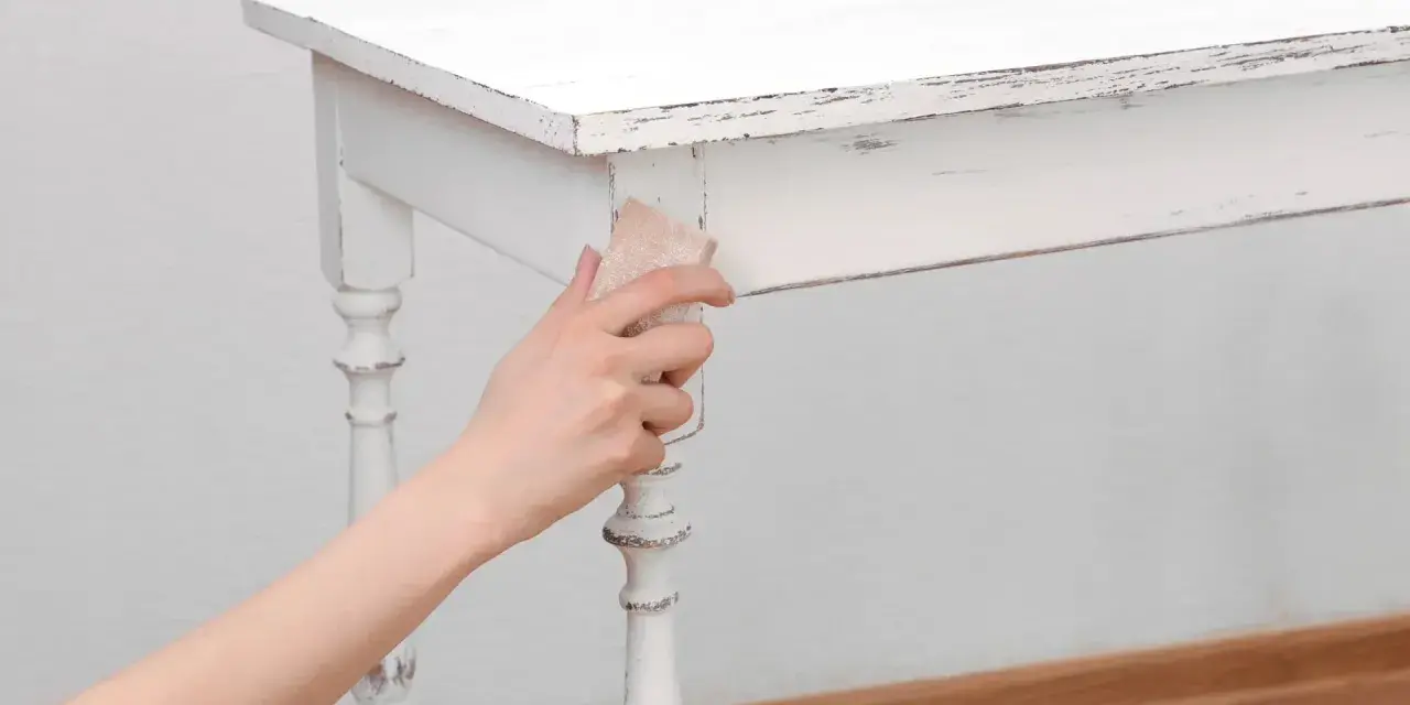 Main tenant enlever peinture sur bois d'une table blanche vieillie, révélant le grain du bois.