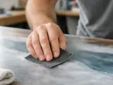 Main d'artisan ponçant une surface pour peindre sur du vernis. La poussière fine indique le travail de préparation.