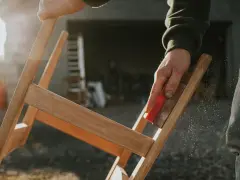 Main d'artisan ponçant un meuble en bois, préparant la surface pour enlever peinture sur bois acétone. Poussière de bois en suspension.