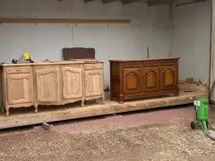 Deux commodes en bois, l'une brute et l'autre vernie, témoignent d'une **réparation meuble en bois**. Un équipement de sablage est prêt à l'emploi.