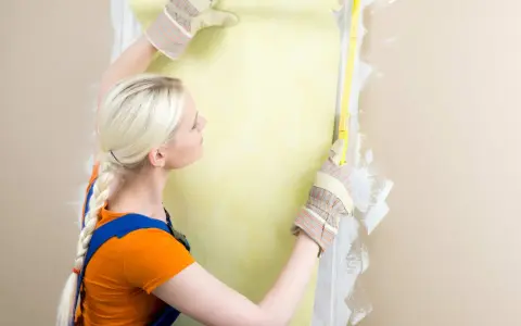 Une femme en salopette bleue et t-shirt orange mesure un mur pour le détapisser puis peindre.