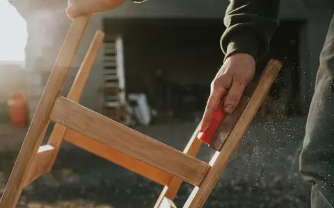 Main d'artisan ponçant un meuble en bois, préparant la surface pour enlever peinture sur bois acétone. Poussière de bois en suspension.