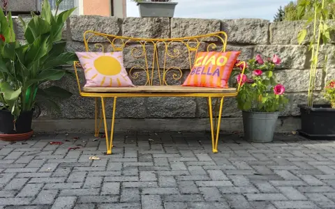 Banc en fer forgé jaune vif, repeint, avec coussins décoratifs. Un soleil rose et "Ciao Bella" sur fond rayé.