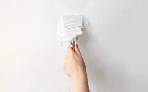 Une main utilise un couteau à mastic pour enlever de la vieille peinture écaillée d'un mur blanc, une étape essentielle pour savoir comment enlever la colle sur un mur avant de peindre.