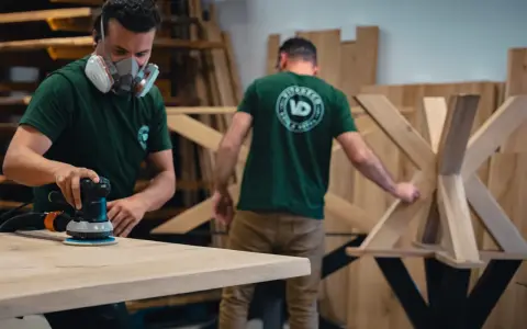 Un artisan, masqué, utilise une ponceuse pour poncer une table en bois. Un autre artisan travaille sur un pied de table en arrière-plan.