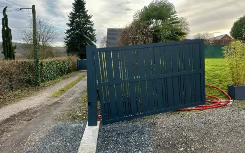 Portail en pente avec gonds sur mesure. Le portail gris foncé est ouvert, révélant un chemin de gravier et une allée bordée de haies.