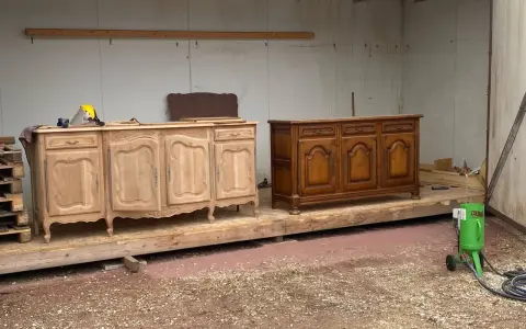 Deux commodes en bois, l'une brute et l'autre vernie, témoignent d'une **réparation meuble en bois**. Un équipement de sablage est prêt à l'emploi.