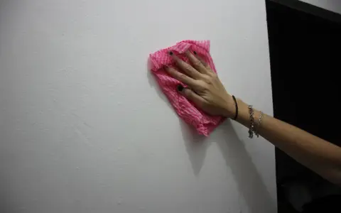Main tenant une lingette rose pour nettoyer un mur. Une astuce pour savoir comment enlever de la colle forte sur un mur.