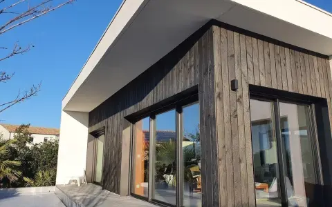 Façade moderne avec bardage en **bois brulé**, grandes baies vitrées reflétant le ciel bleu et la végétation.