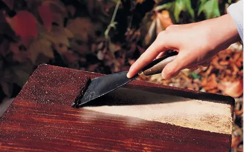 Main d'une personne utilisant un grattoir pour enlever de la peinture d'un meuble en bois, comme avec un décapant peinture bois fait maison.