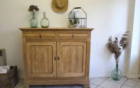 Buffet en bois clair, prêt pour sa restauration. Deux tiroirs et deux portes ornées de moulures. Décoration champêtre avec des vases anciens et des herbes sèches.