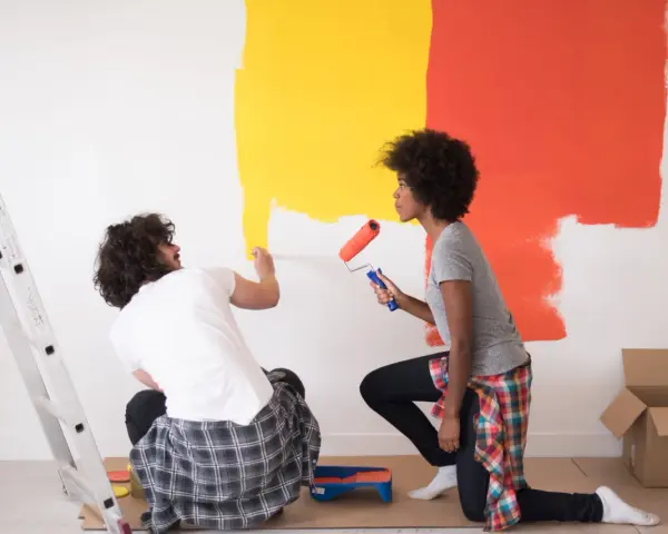 Un couple peint un mur en jaune et orange. Ils utilisent des rouleaux pour nettoyer peinture glycero. Une échelle est à gauche.